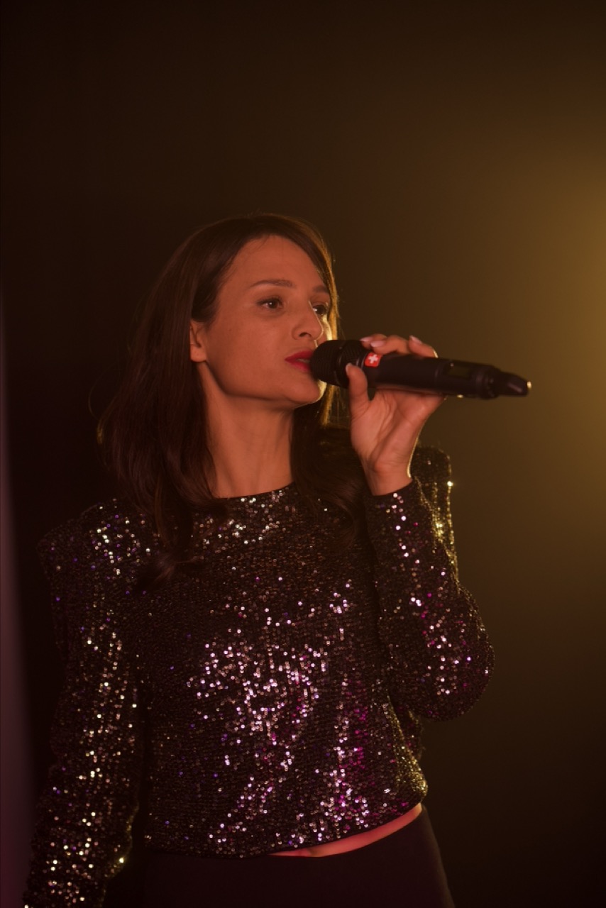Woman in a black sequin top singing into a microphone with warm stage lighting, perfect as your wedding singer for an unforgettable celebration.