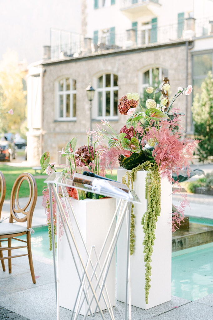 Modernes Outdoor-Hochzeitssetup in der Schweiz mit hohen weißen Blumenarrangements, Stühlen und einer professionellen Traurednerin nahe einem eleganten Gebäude.