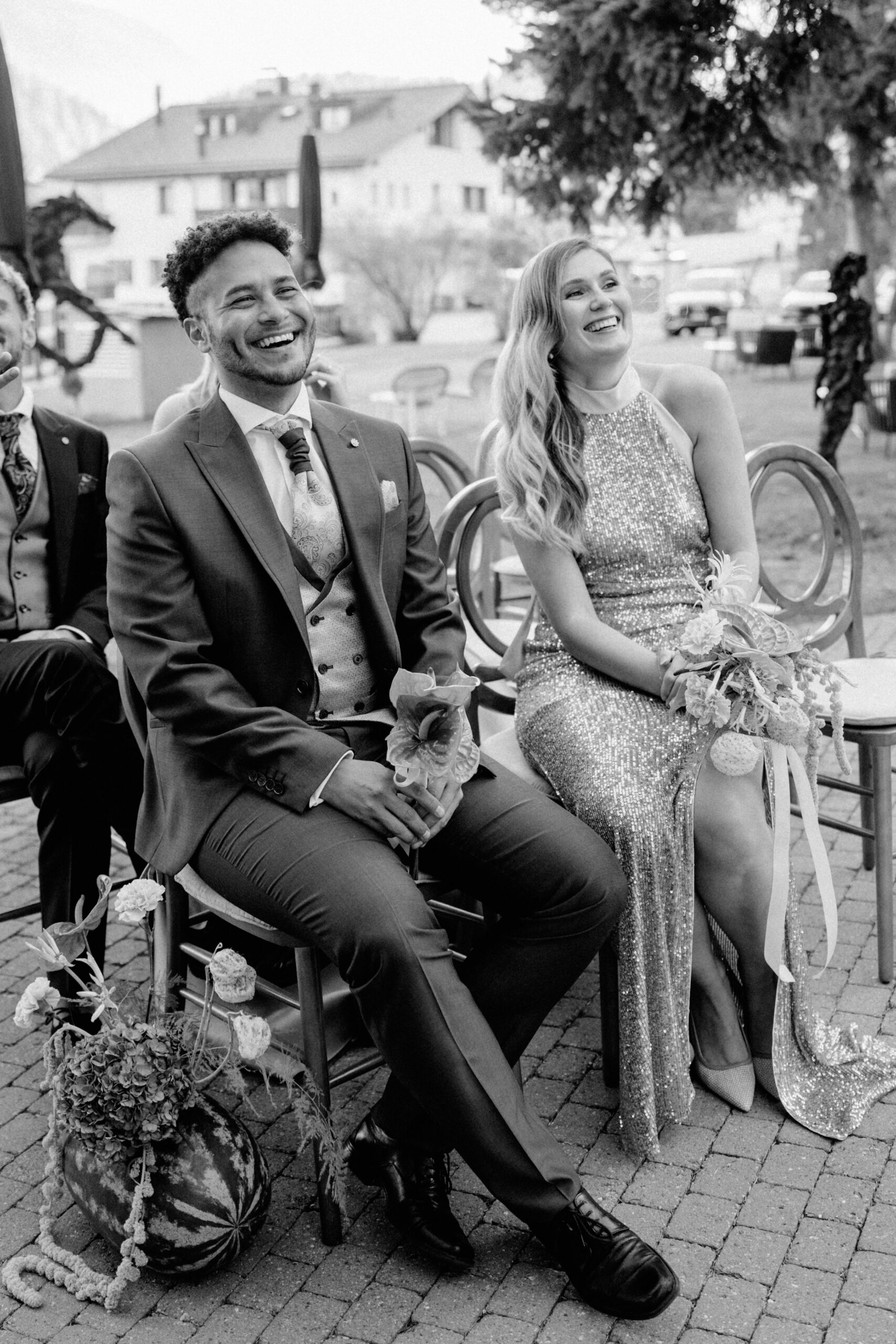 Ein lächelnder Mann in einem Anzug und eine Frau in einem glitzernden Kleid sitzen und lachen bei einer Veranstaltung im Freien in Zürich.