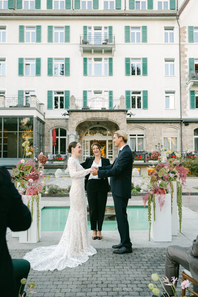 Bride and groom hold hands during an outdoor wedding ceremony in front of a hotel, while their officiant presides behind them.
