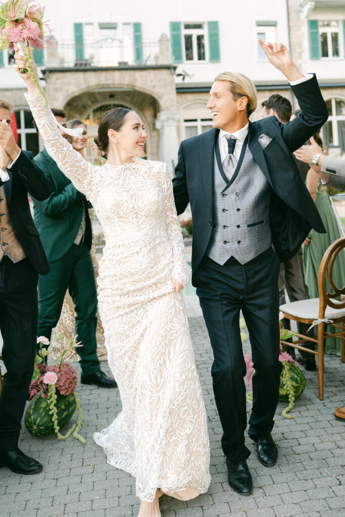 Ein Brautpaar feiert fröhlich unter freiem Himmel in Zürich, hebt die Arme und lächelt inmitten der Gäste, während ein Hochzeits-DJ und eine Hochzeitssängerin für die perfekte Hochzeitsatmosphäre sorgen.