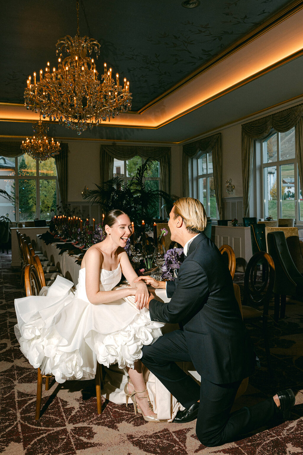 Bride and groom laugh together at an elegantly decorated reception table under chandeliers, while their wedding DJ in Switzerland creates the perfect atmosphere for an unforgettable evening.