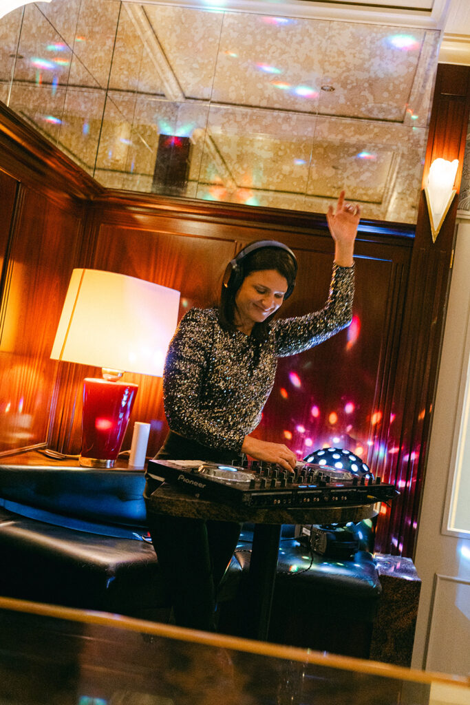 A wedding DJ wearing headphones and a sparkly top plays music and raises one hand in a warmly lit room with disco lights, bringing vibrant energy to the festivities in Zurich, Switzerland.