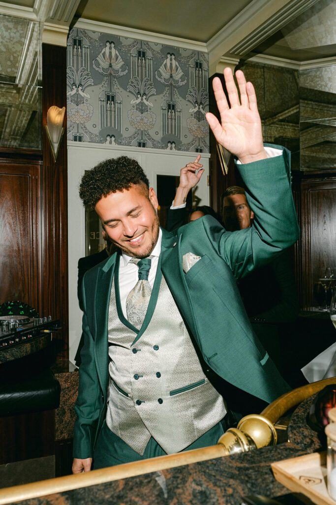 Ein Mann in einem grünen Anzug lächelt und hebt die Hand, während er an einer Bar in einem eleganten Raum in Zürich steht.