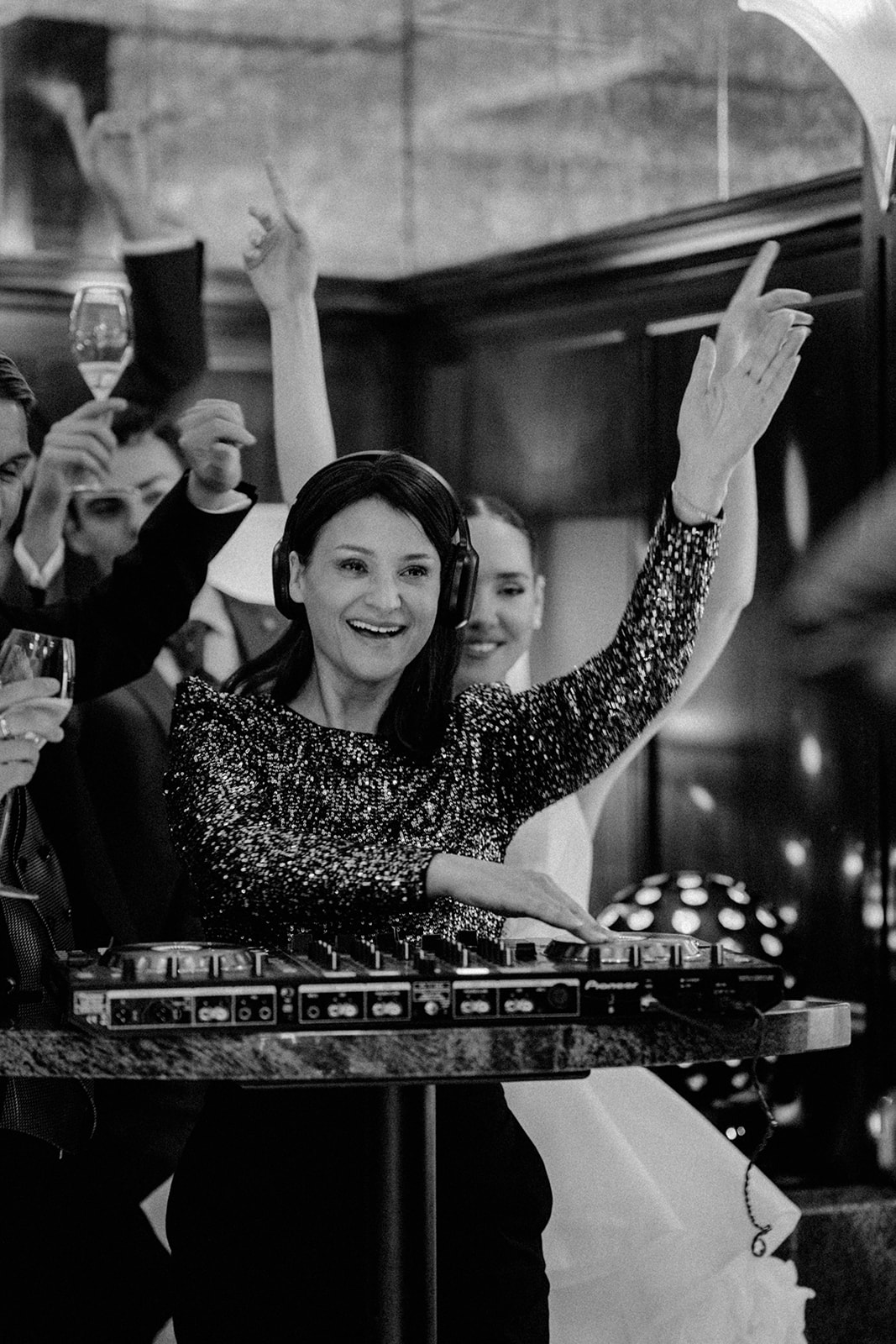 Frau DJ in paillettenbesetztem Oberteil lächelnd und mit erhobener Hand, umgeben von tanzenden Menschen auf einer lebhaften Party in der Schweiz-perfekt für jede Hochzeit mit einer talentierten Hochzeitssängerin oder Traurednerin.