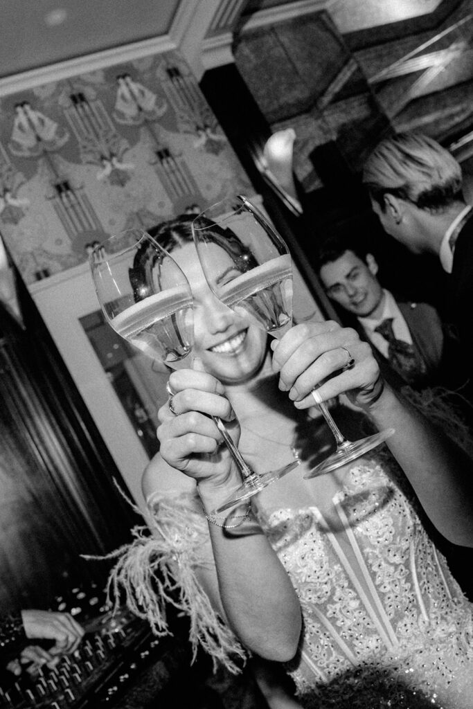 Eine Frau lächelt und hält zwei Weingläser vor ihr Gesicht bei einer lebhaften Indoor-Party in Zürich, wo sie mit Freunden feiert.