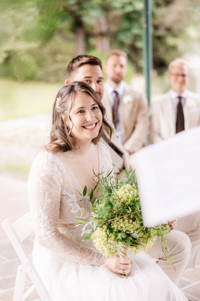 A smiling bride in a lace dress holds a bouquet of flowers while the bridesmaids sit in the background at a wedding in Zurich, Switzerland, ready to celebrate with the perfect wedding DJ.