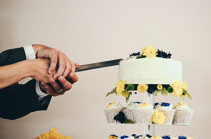 Zwei Hände schneiden eine weiße, mit gelben und blauen Blumen verzierte Torte an, darüber passende Cupcakes - ein eleganter Moment in Zürich, perfekt für einen Hochzeits-DJ oder eine Traurednerin zum Mitfeiern.