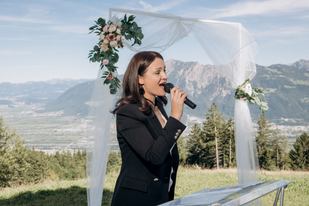 Traurednerin in schwarzem Blazer spricht mit Mikrofon im Freien, vor atemberaubender Bergkulisse und Hochzeitsbogen in der Schweiz.