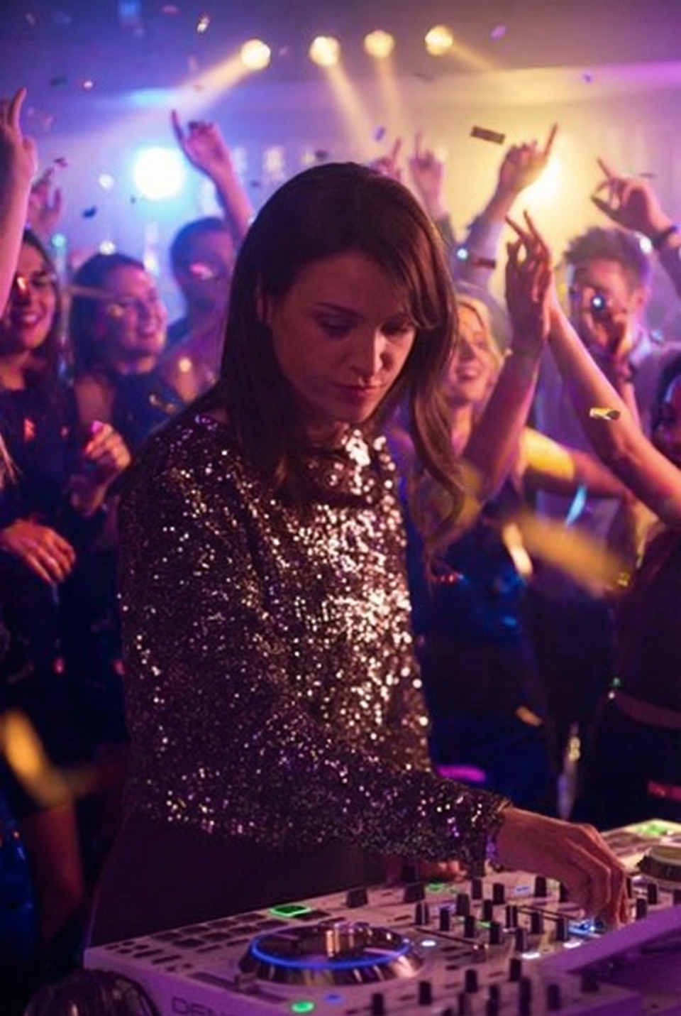 Ein dynamischer DJ in einem glitzernden Oberteil heizt der ausgelassenen Party richtig ein, während er Musik auflegt, die Leute tanzen und überall Konfetti herabregnet.